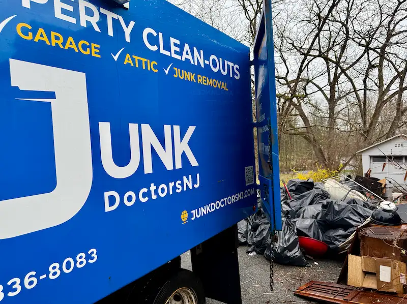 A Junk Doctors NJ hauling truck parked next to a massive pile of bagged garbage, an old grill, and wood debris removed during a backyard shed cleanout.