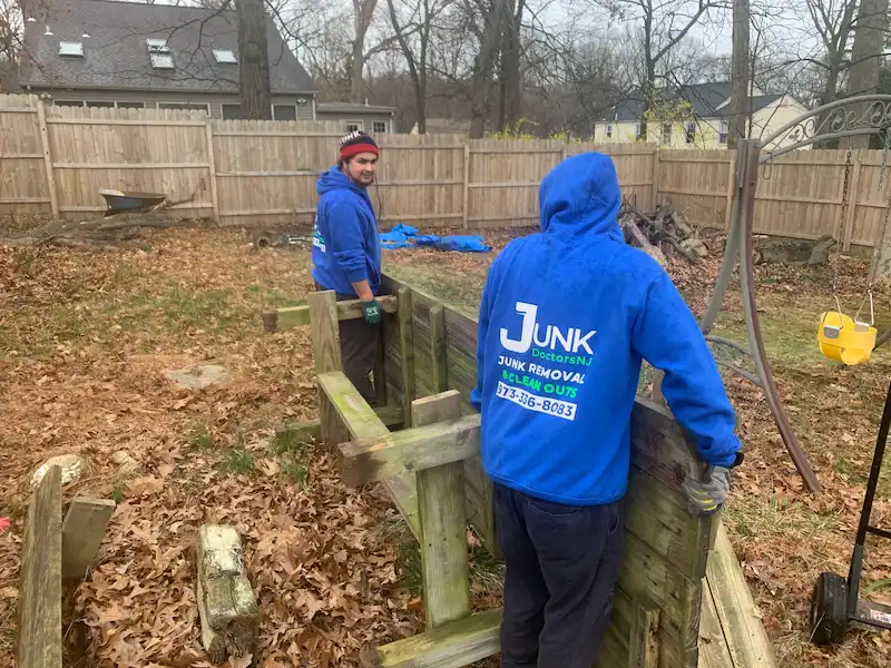 Two JunkDoctors NJ crew members lifting a heavy, weathered wooden picnic table and other yard items during a residential backyard cleanup service.