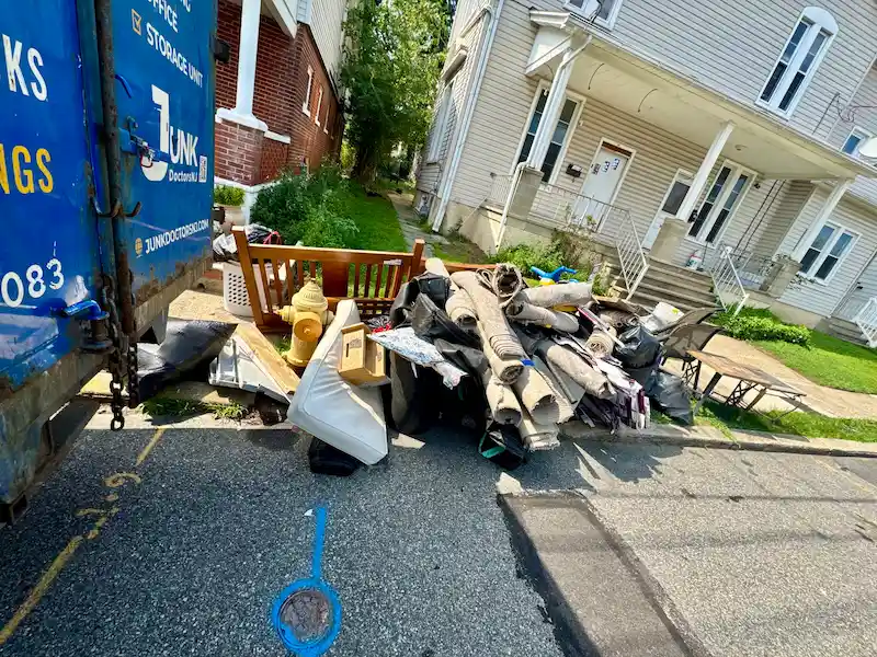 A variety of household junk including rolls of carpet, furniture, and a mattress staged for curbside pickup by JunkDoctors NJ in a North Jersey neighborhood.