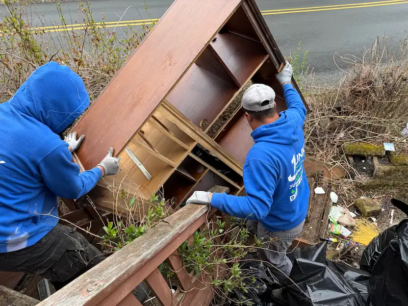Two professional Junk Doctors crew members in blue uniforms safely carrying a heavy wooden entertainment center down a set of outdoor wooden stairs.