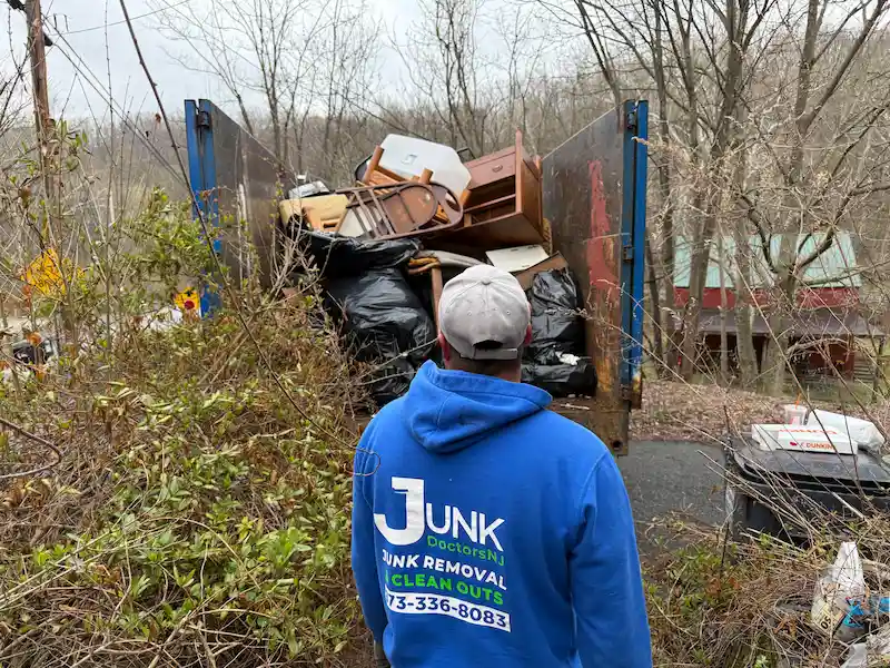 A JunkDoctors NJ worker standing on a litter-strewn path, looking toward a blue-sided dump truck that is completely full and heavily loaded with discarded furniture, storage bins, trash bags, and other junk.