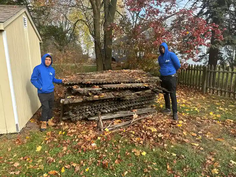 Two JunkDoctors team members in blue hoodies standing with a large stack of old wood fencing and yard debris in a New Jersey backyard.
