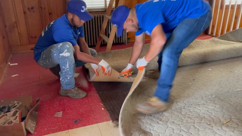 Junk Doctors NJ team using safety gear and tools to cut and roll up old carpet for efficient removal from a home.
