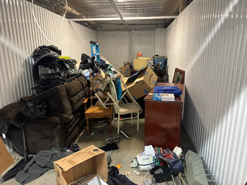Before shot of a cluttered storage unit in North NJ filled with an old recliner, wooden chairs, and moving boxes.